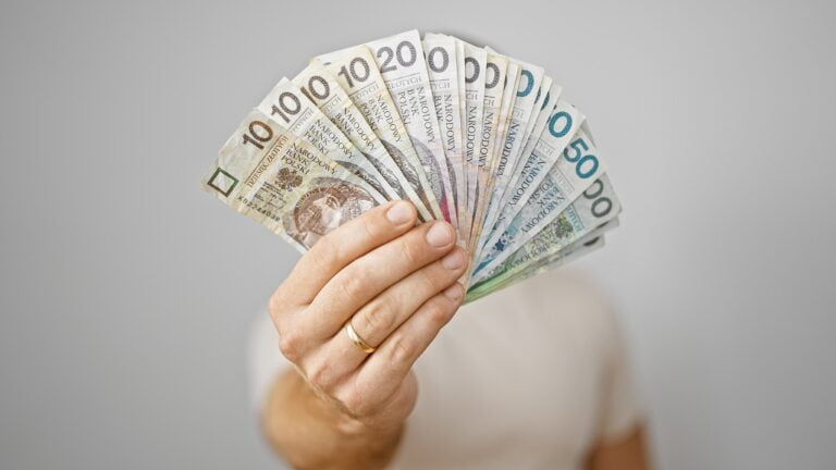 Dłoń trzymająca wachlarz polskich banknotów o różnych nominałach na jednolitym tle.