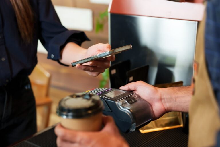 Klientka płacąca smartfonem przy terminalu płatniczym, w tle widoczny trzymający kawę barista.