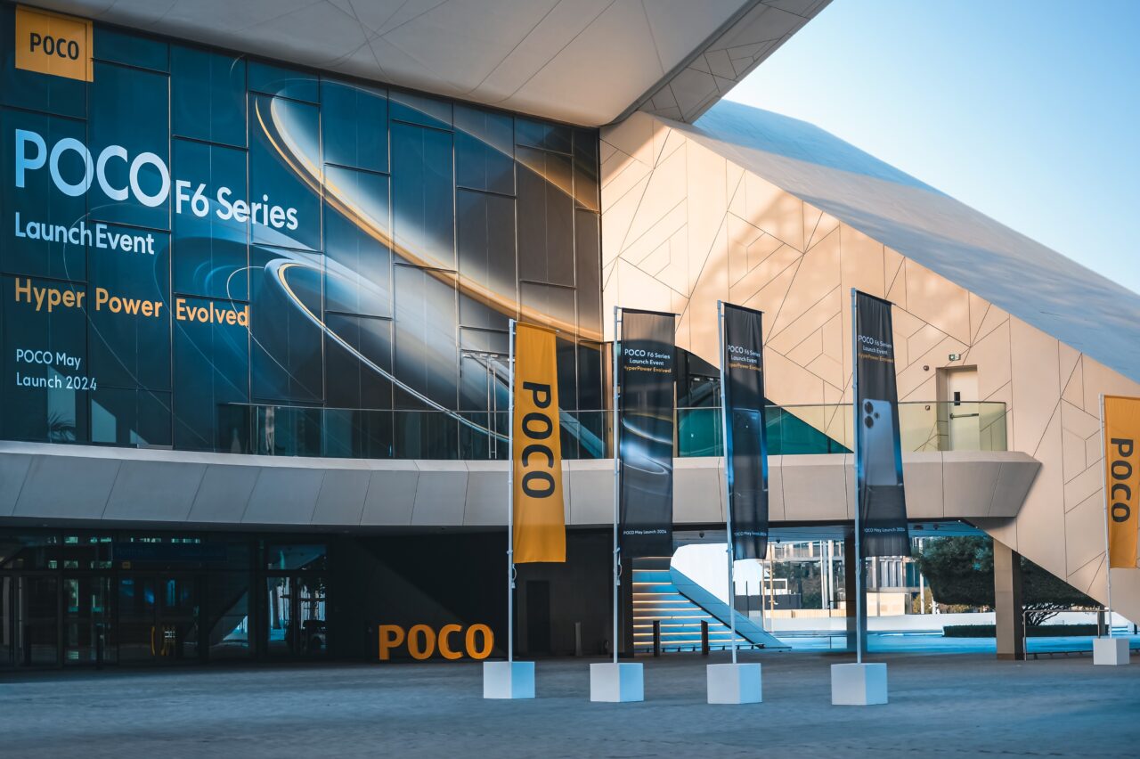 Zewnętrze budynku z dużym banerem promującym premierę serii POCO F6 planowaną na maj 2024, z hasłami "Hyper Power Evolved" i flagami z logo POCO.