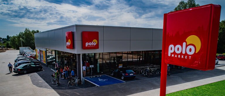 Sklep PoloMarket z logo na dużym czerwonym znaku i na budynku, zaparkowane samochody, ludzie wchodzący do sklepu, parking dla niepełnosprawnych.