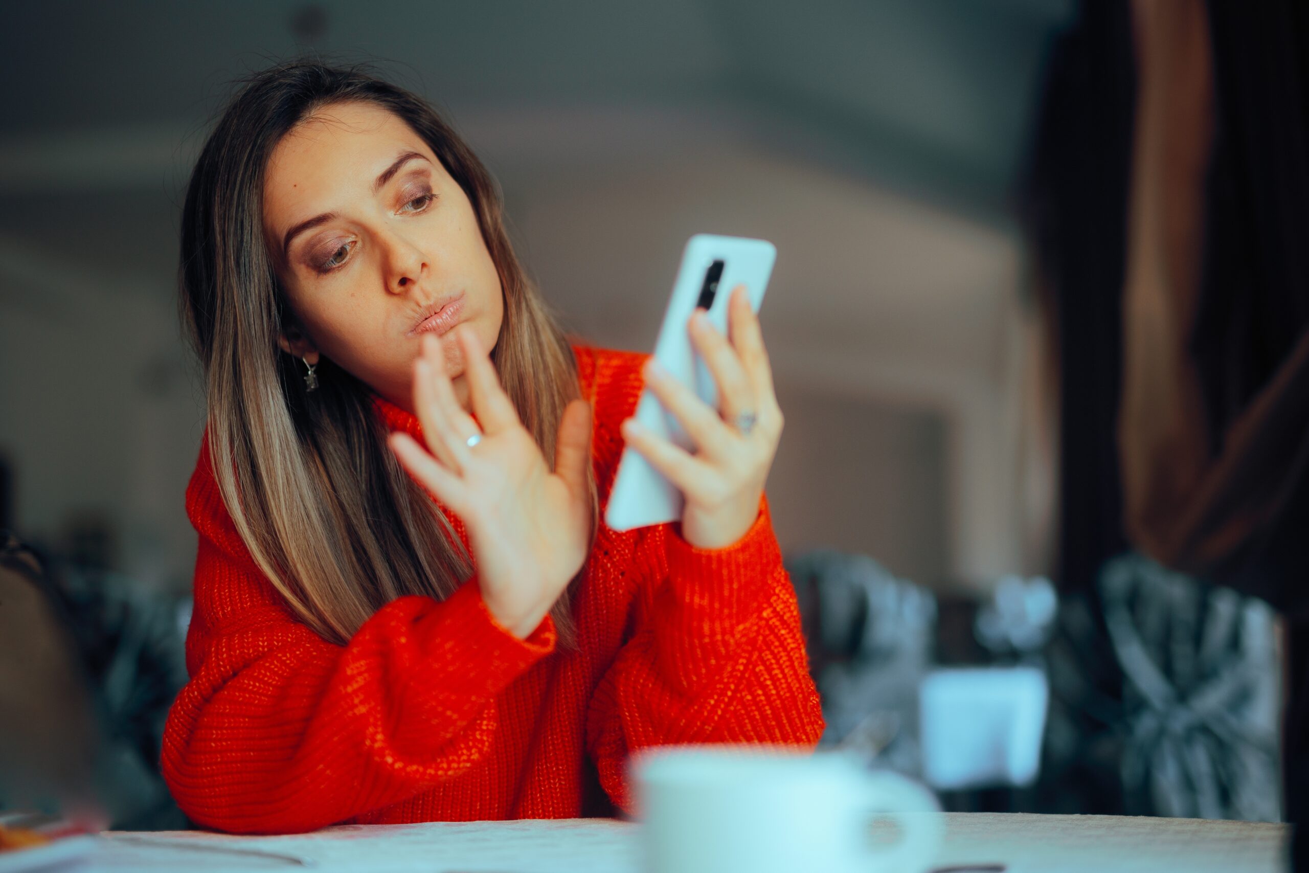 Kobieta w czerwonym swetrze, patrząca na telefon komórkowy i dotykająca swojej twarzy palcem wskazującym.