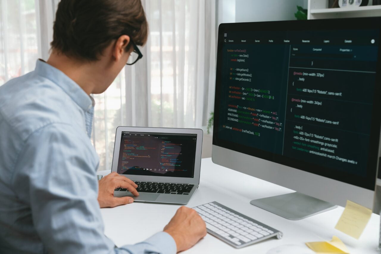 Programista pracujący przy komputerze z kodem wyświetlanym na monitorze oraz laptopie.
