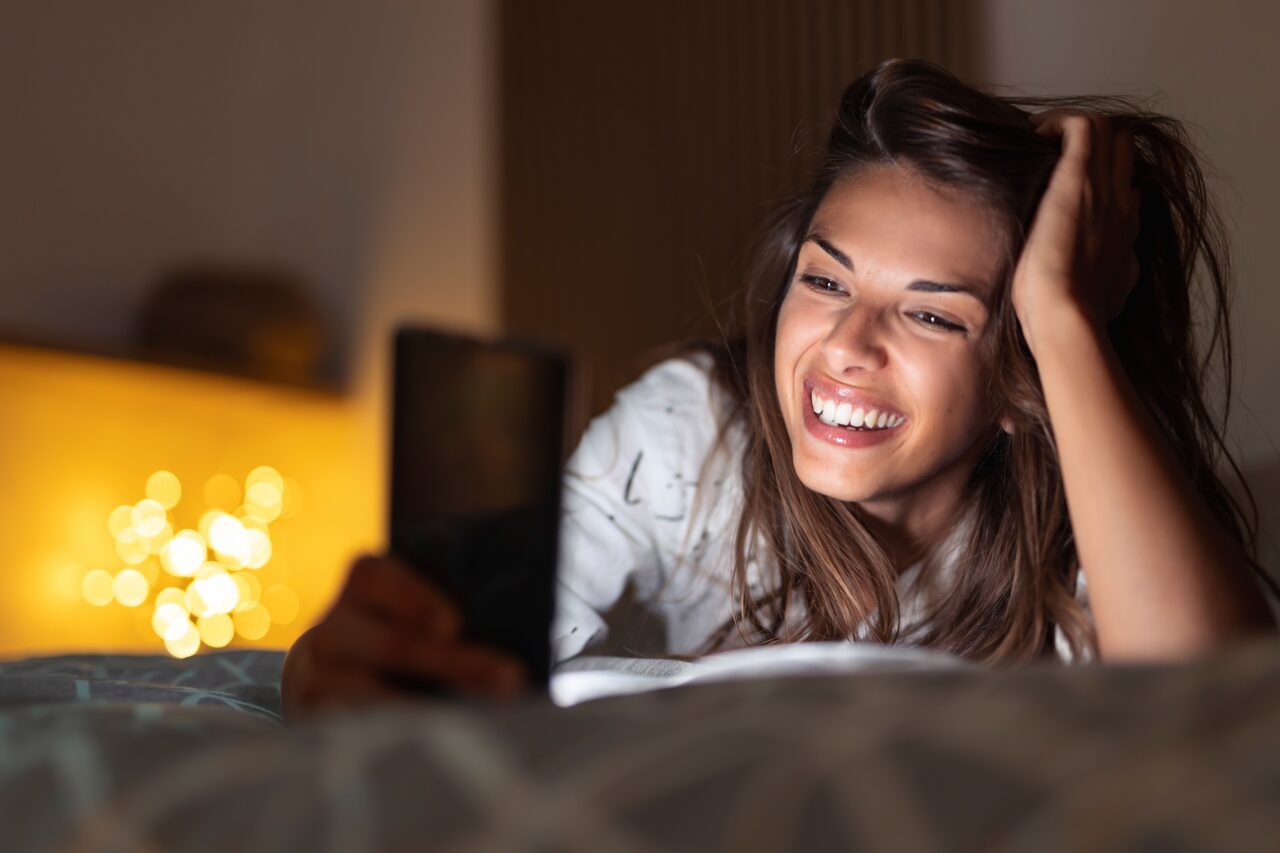 Kobieta uśmiechająca się, leżąca na łóżku, patrząca na telefon. Portale randkowe to dla niej codzienność