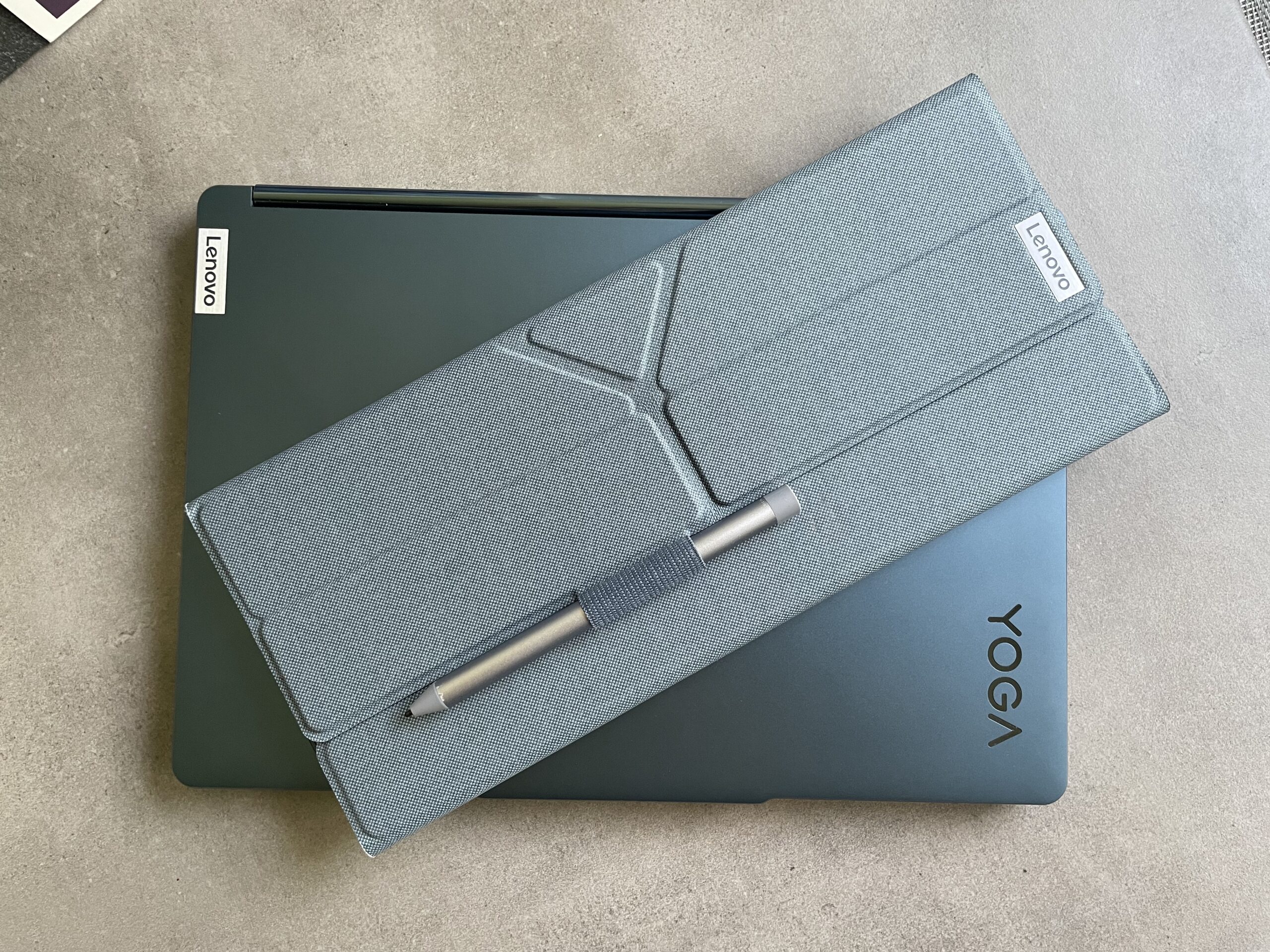 Laptop Lenovo Yoga w niebieskiej obudowie z etui w kolorze szarym i rysikiem na nim położonym.