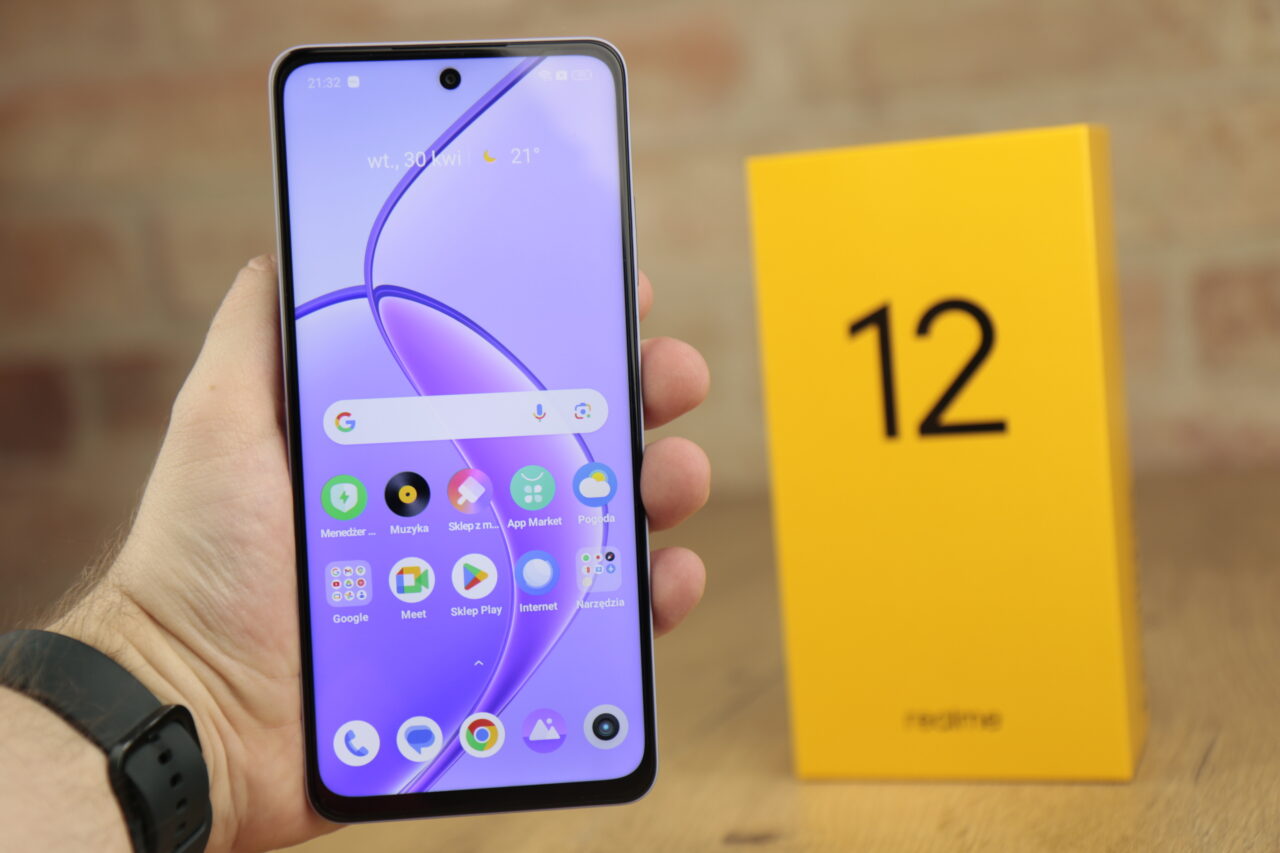 Recenzując realme 12 5G, telefon trzymany jest w dłoni, pokazując przedni ekran z wyświetloną aplikacją oraz żółte opakowanie z numerem 12.