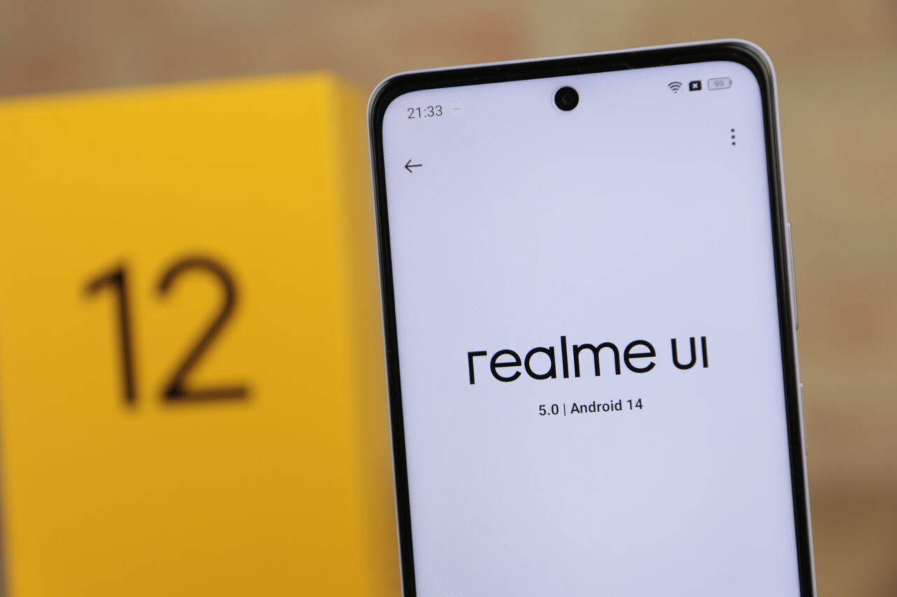 Recenzowany smartfon realme 12 5G z ekranem pokazującym logo "realme UI" i informację o wersji systemu Android 14, na tle żółtego elementu z numerem 12.