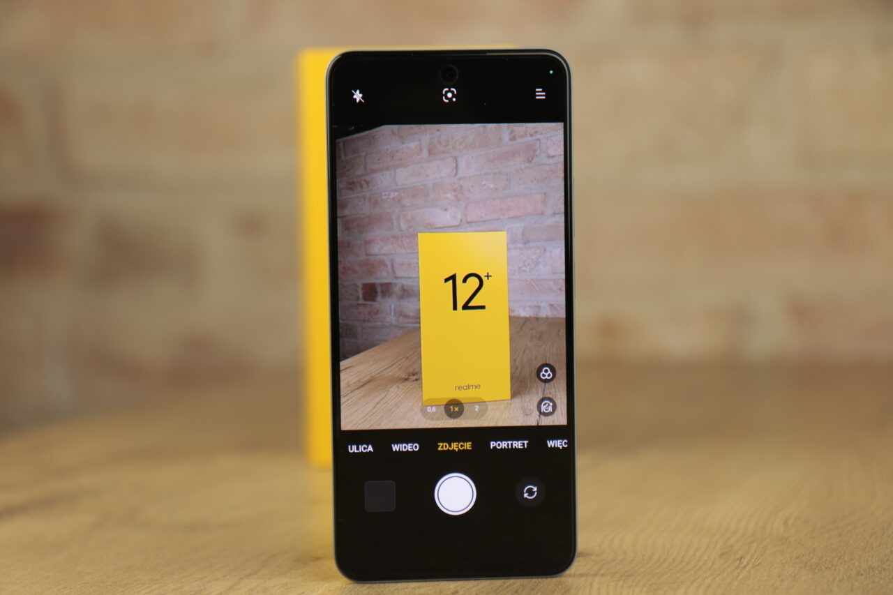 Smartfon realme 12+ 5G leżący na drewnianym stole, wyświetlający aplikację kamery z widokiem na żółtą kartkę z napisem 12+ oraz realme. Recenzja realme 12+ 5G.