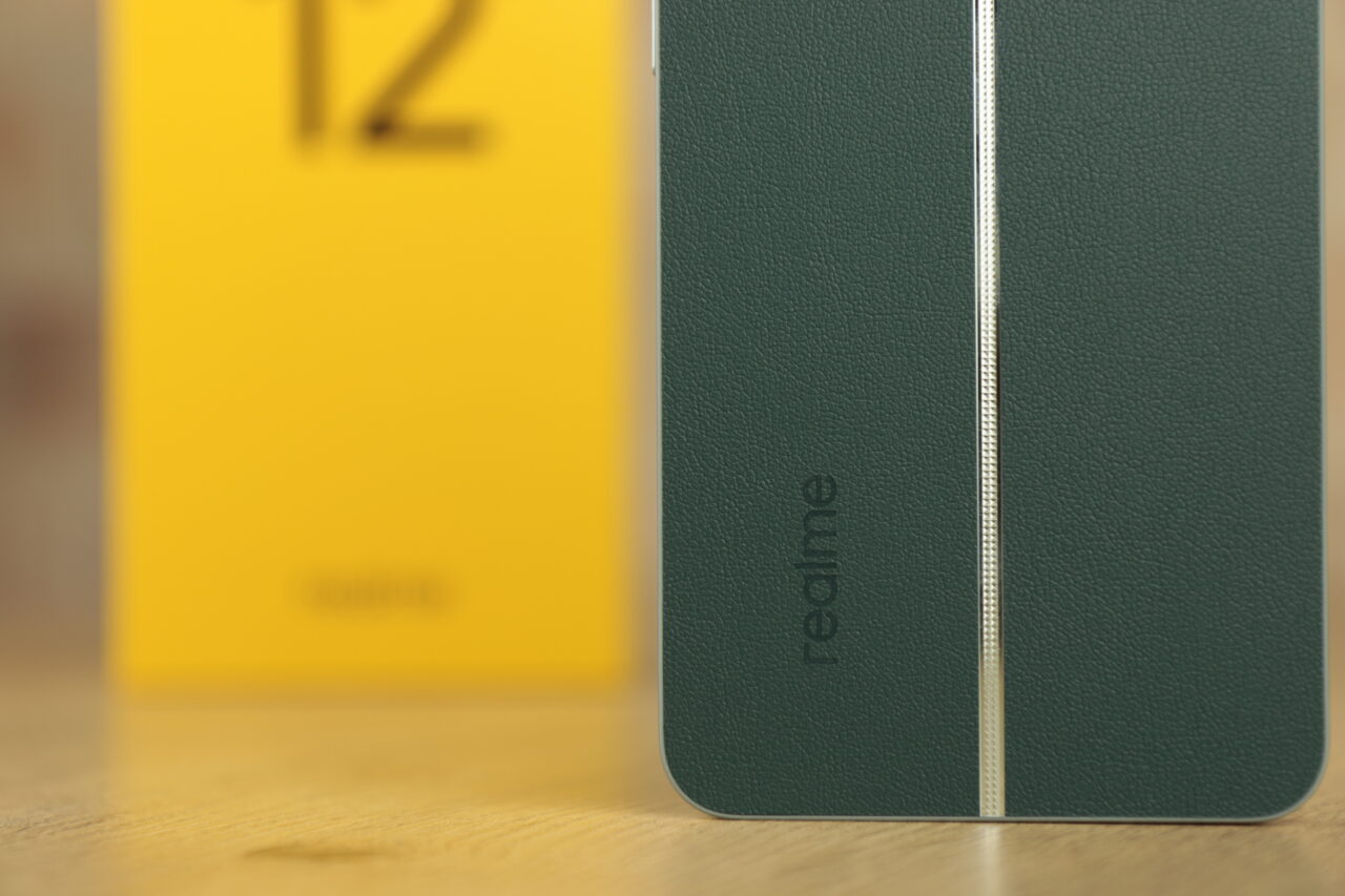 Zbliżenie na zielone etui telefonu Realme 12+ 5G z białą linią dekoracyjną i nazwą marki, nieostry żółty pudełko w tle.