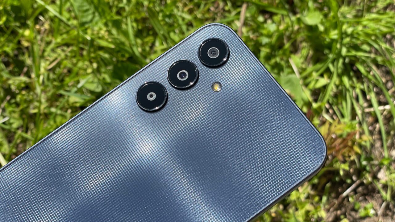 Smartfon leżący na trawie, z wyeksponowanymi trzema apararami i lampą błyskową na niebieskim tle z fakturą.