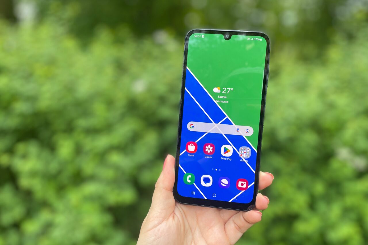 Telefony na komunię. Dłoń trzymająca smartfon z ekranem pokazującym aplikacje i temperaturę w Warszawie na zielonym tle roślinności.