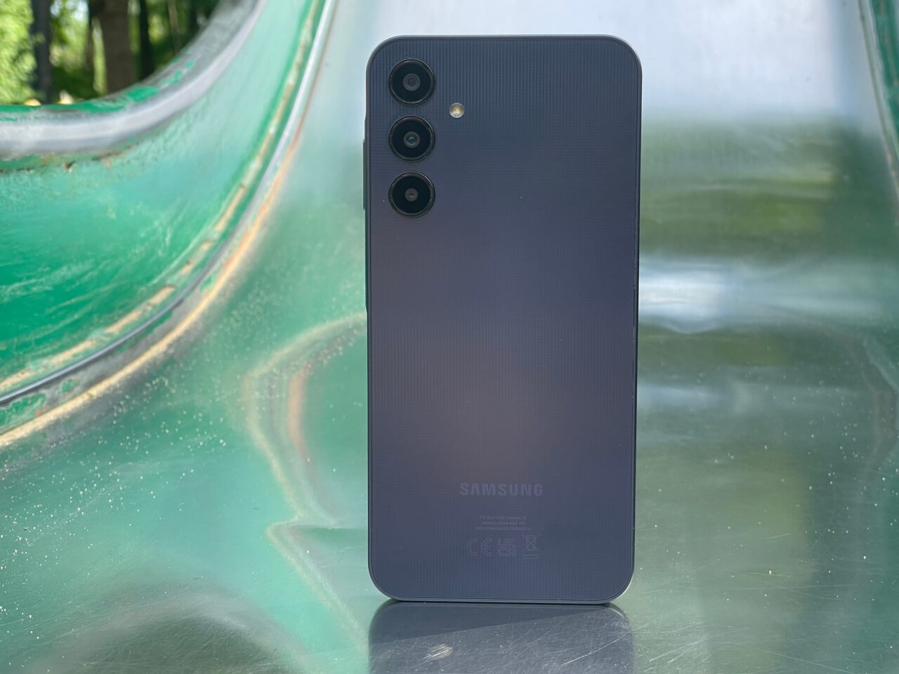 Szary smartfon Samsung stoi na szklanej powierzchni z zielonym tłem.