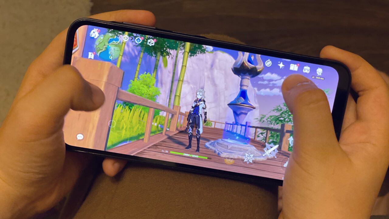Osoba trzyma smartfon, na którym wyświetlana jest gra z postacią stojącą na drewnianym mostku w fikcyjnym świecie.