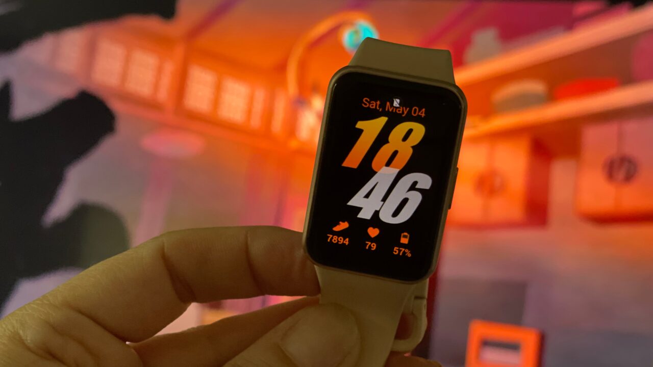 Ręka trzymająca inteligentny zegarek z wyświetlaczem pokazującym godzinę 18:46 i datę (sobota, 4 maja), liczbę kroków, tętno i poziom baterii, w tle rozmyte wnętrze o ciepłej kolorystyce.