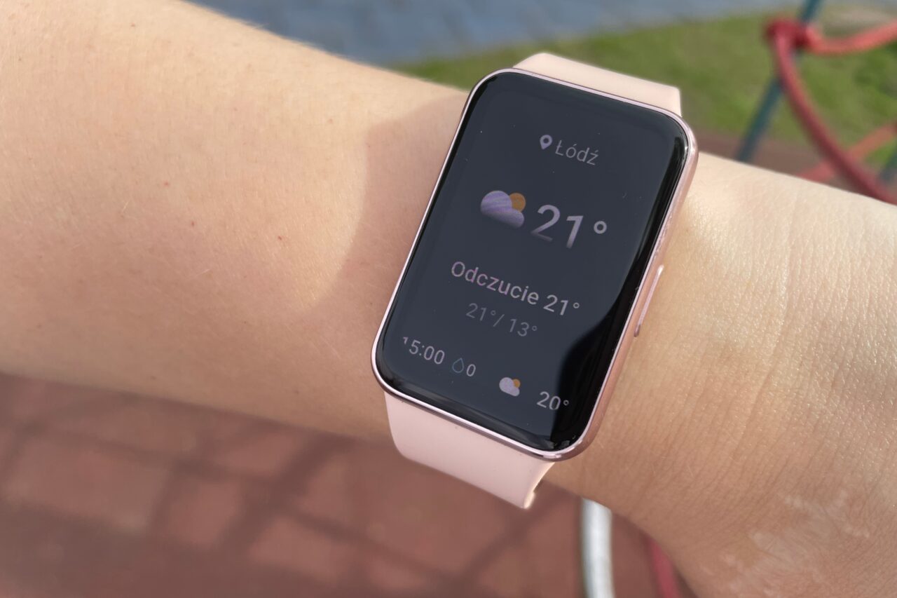 Różowy smartwatch na nadgarstku pokazujący pogodę dla Łodzi z temperaturą 21°C.
