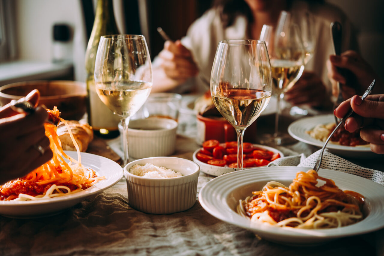 Osoby jedzące spaghetti przy stole, na którym znajdują się kieliszki z białym winem, miseczki z serem i pokrojonymi pomidorami.