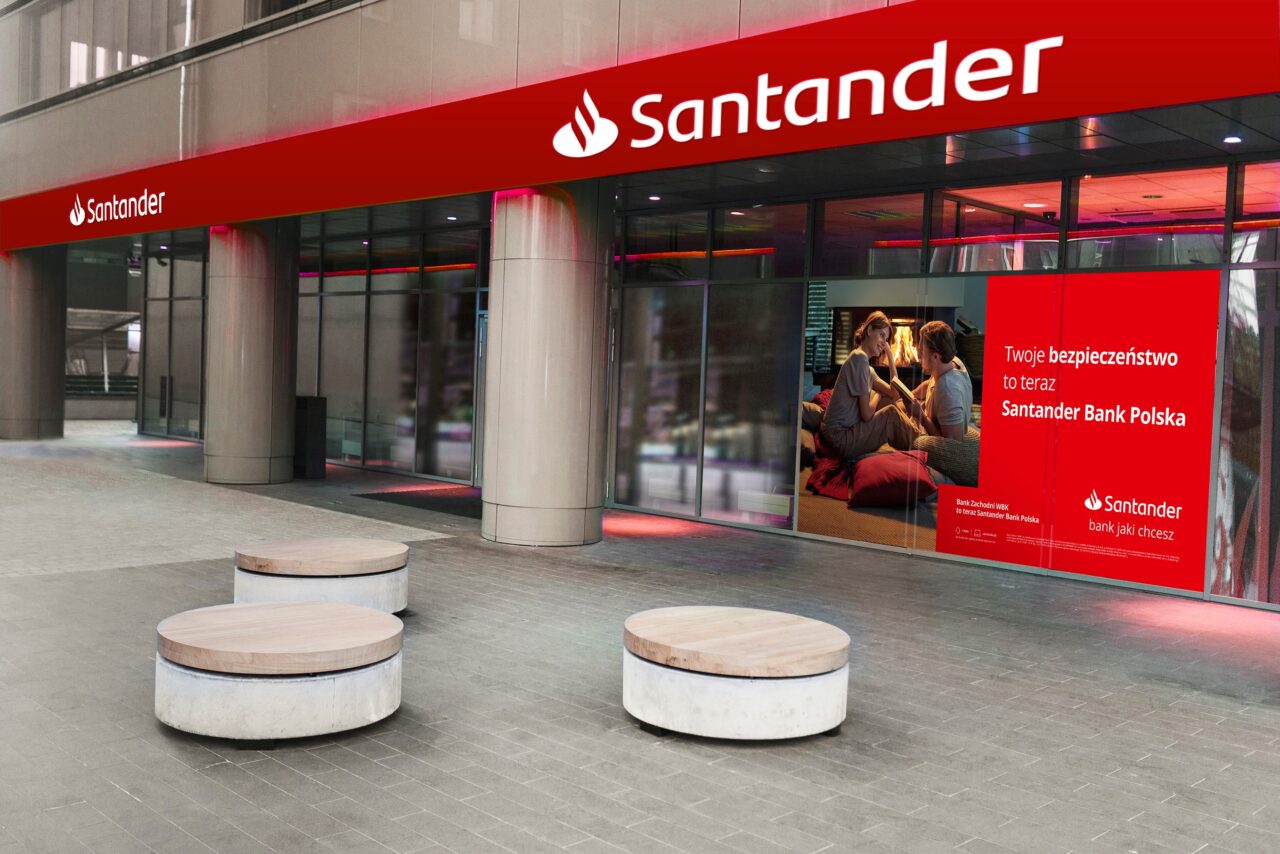 Wejście do banku Santander z czerwonym szyldem oraz plakatem reklamującym bank. Przed budynkiem znajdują się okrągłe siedziska.