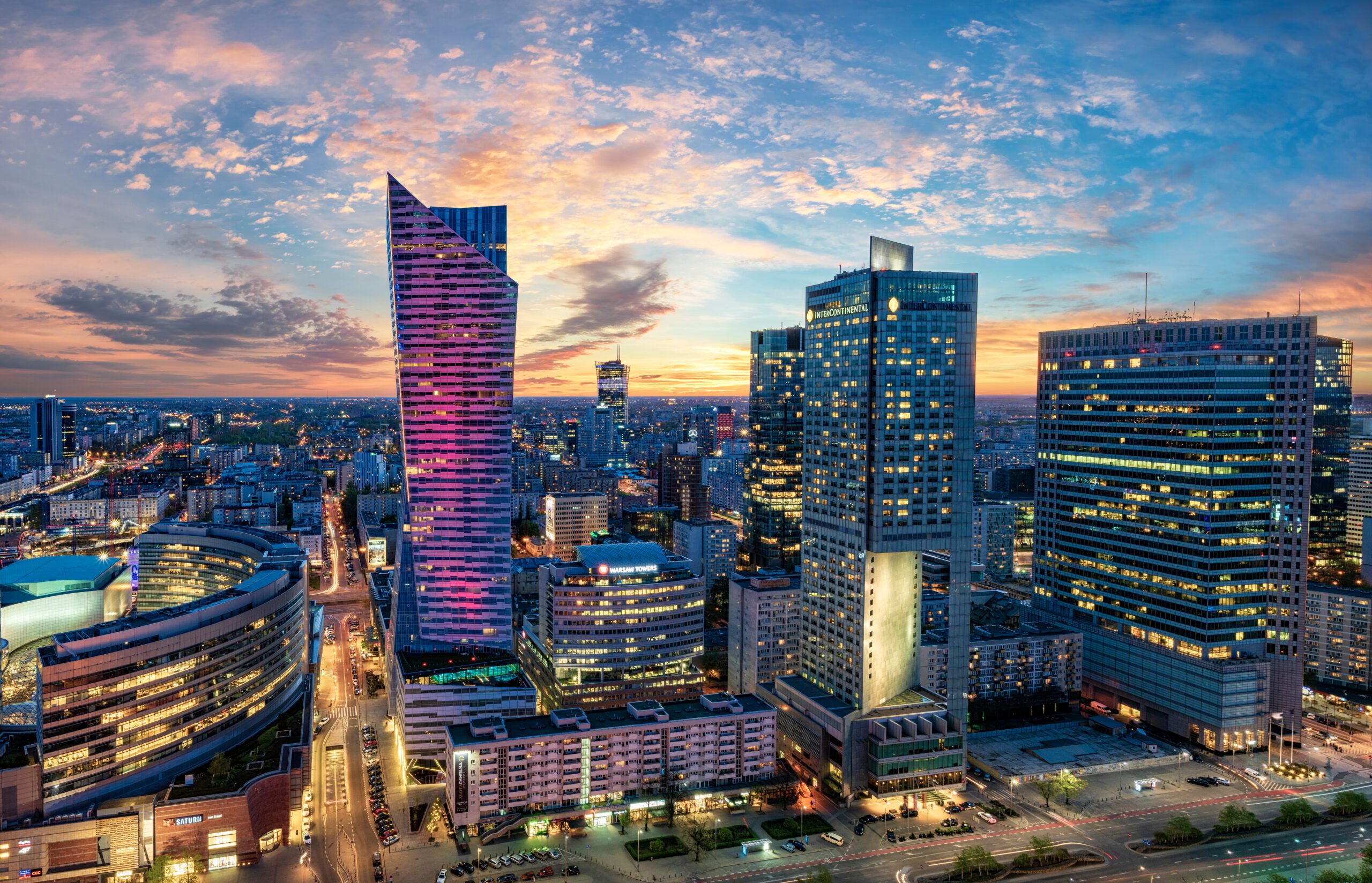 Panorama miasta z wieżowcami w tle o zmierzchu, jasne, kolorowe światła na budynkach, niebo w różowo-niebieskich barwach.