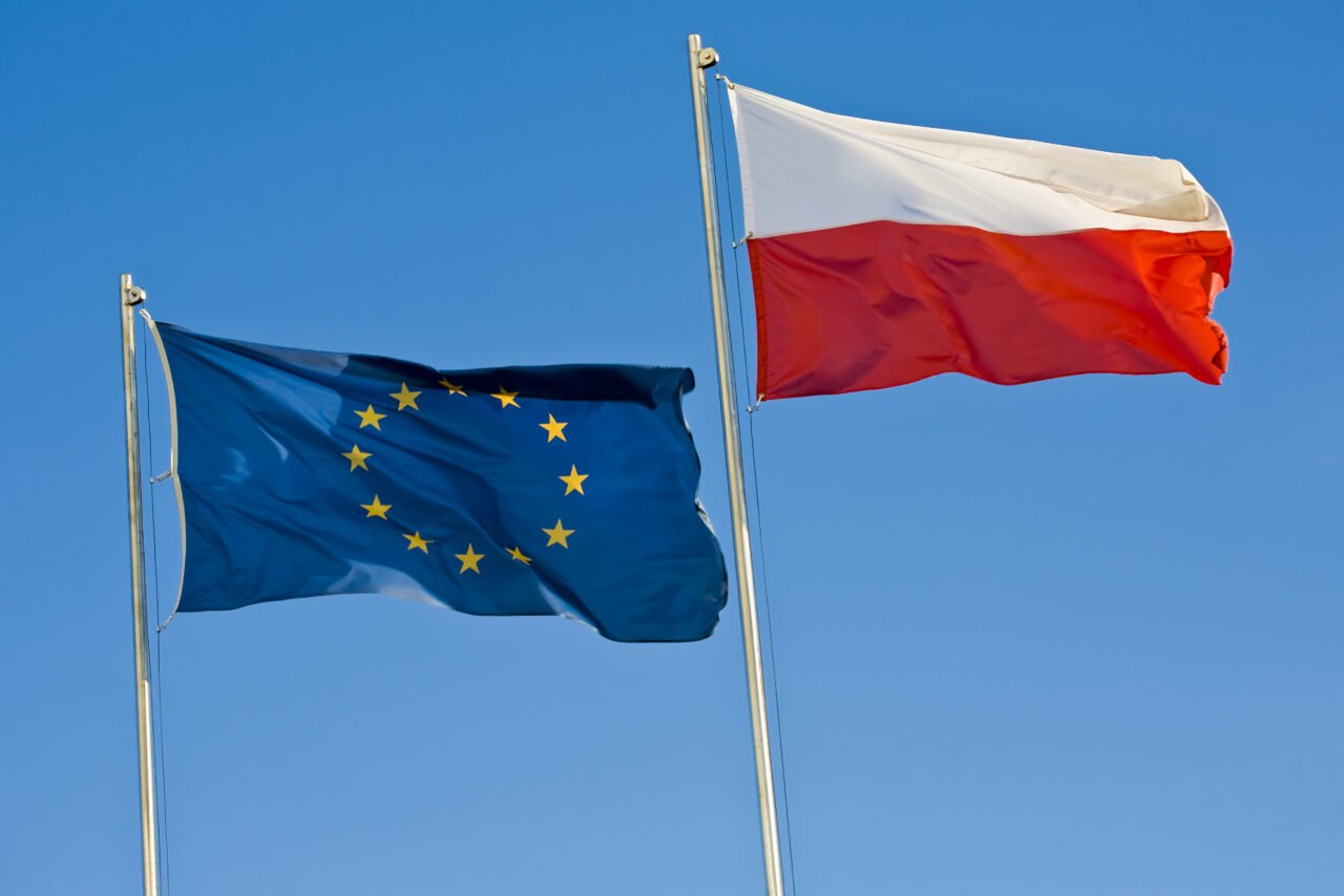 Flagi Unii Europejskiej i Polski powiewające na masztach na tle niebieskiego nieba.