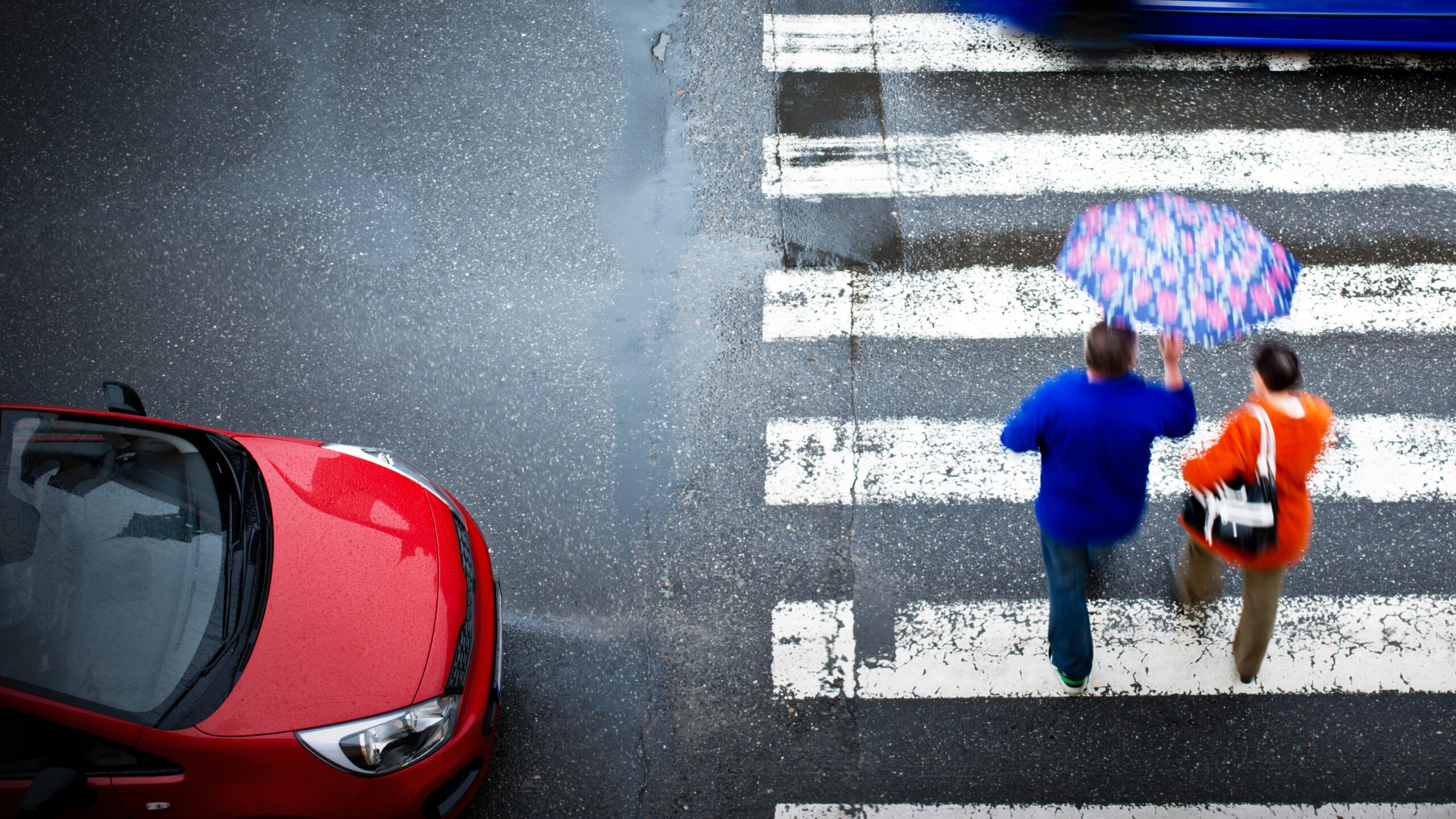 Widok z góry na przejście dla pieszych w deszczu, gdzie dwóch ludzi w kurtkach przechodzi przez pasy trzymając kolorowy parasol, a obok nich stoi czerwony samochód.