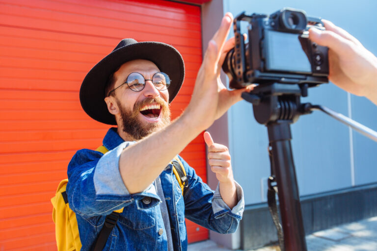 Mężczyzna z okularami i czarnym kapeluszem uśmiecha się i pokazuje kciuk do kamery filmowej na tle czerwonej ściany.