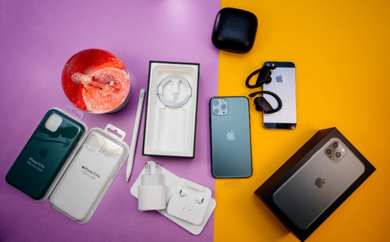Produkty Apple, w tym iPhone, etui, ładowarki, słuchawki, rysik oraz stojący na stole lampion, na tle żółto-fioletowej powierzchni.