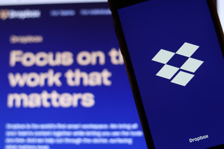 Telefon komórkowy z logotypem Dropbox na wyświetlaczu, w tle monitor komputerowy z rozmytym tekstem "Focus on the work that matters".