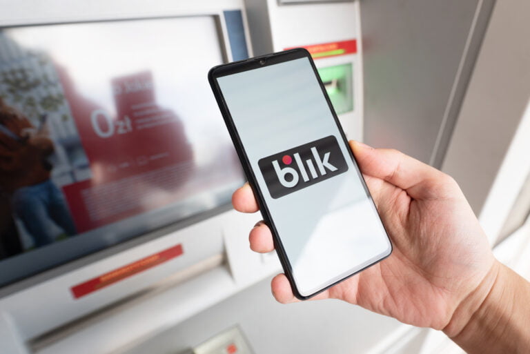 Dłoń trzymająca smartfon z logo BLIK na ekranie przed bankomatem.
