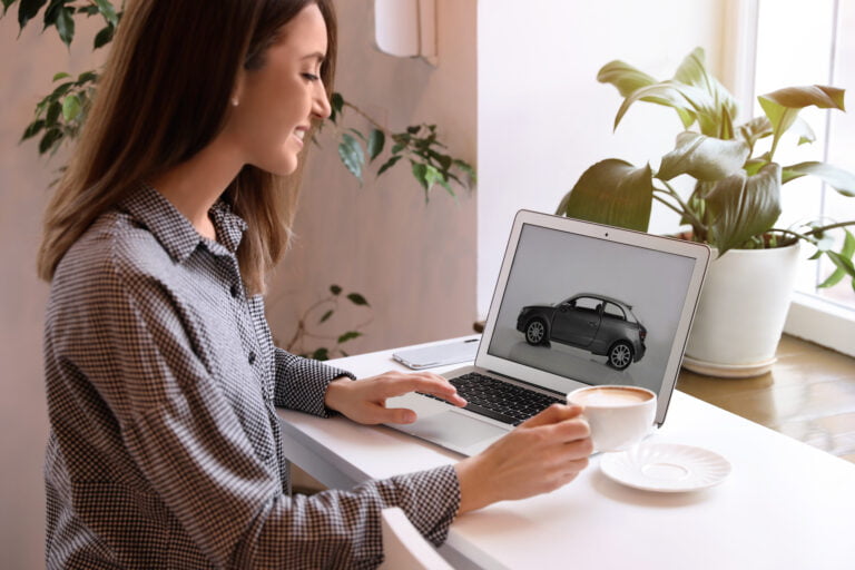 Kobieta, która używa laptopa z obrazem samochodu na ekranie, siedzi przy biurku i pije kawę, otoczona roślinami doniczkowymi.