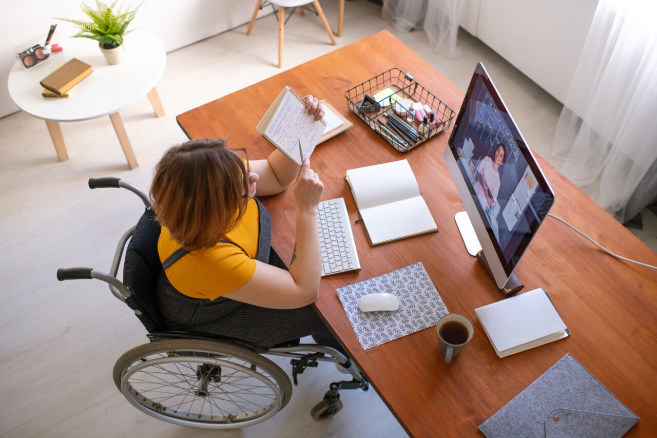 Osoba na wózku inwalidzkim uczestnicząca w wideokonferencji, siedząca przy biurku z drewnianym blatem, na którym leżą zeszyty, klawiatura, myszka, kawa i inne przybory.