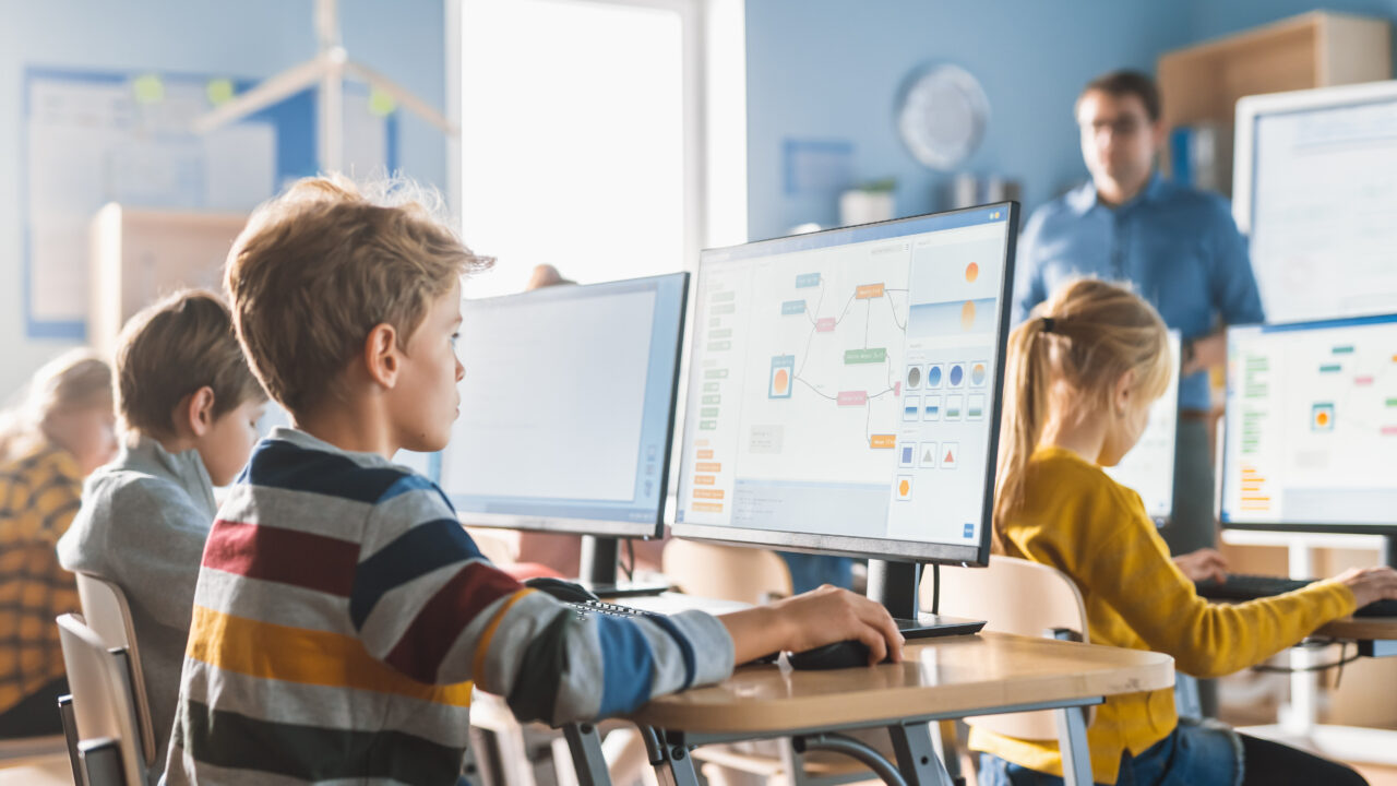 Dzieci uczące się przy komputerach w klasie szkolnej, nauczyciel stojący w tle.
