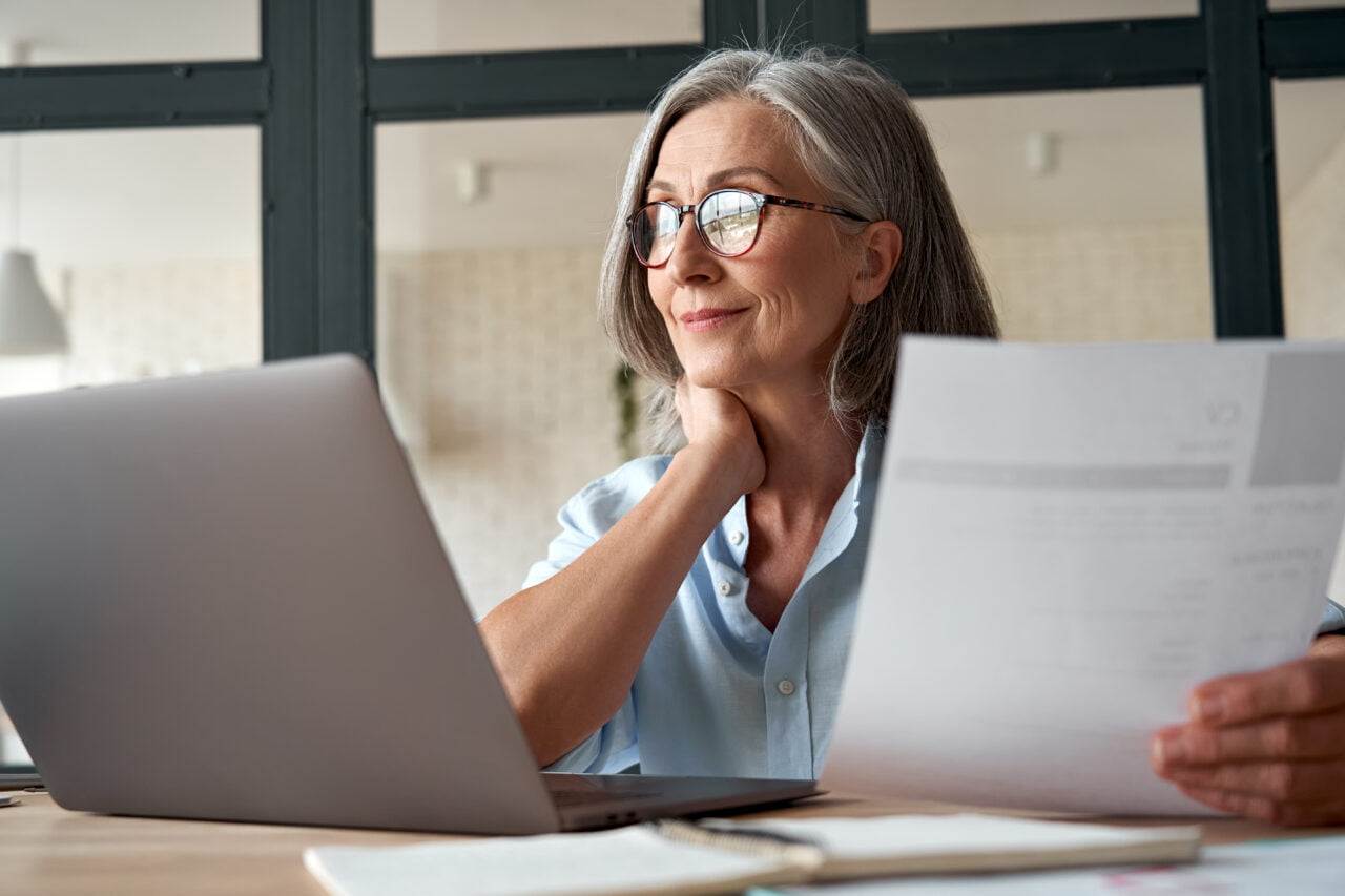 Kobieta z okularami czytająca dokumenty i pracująca na laptopie.