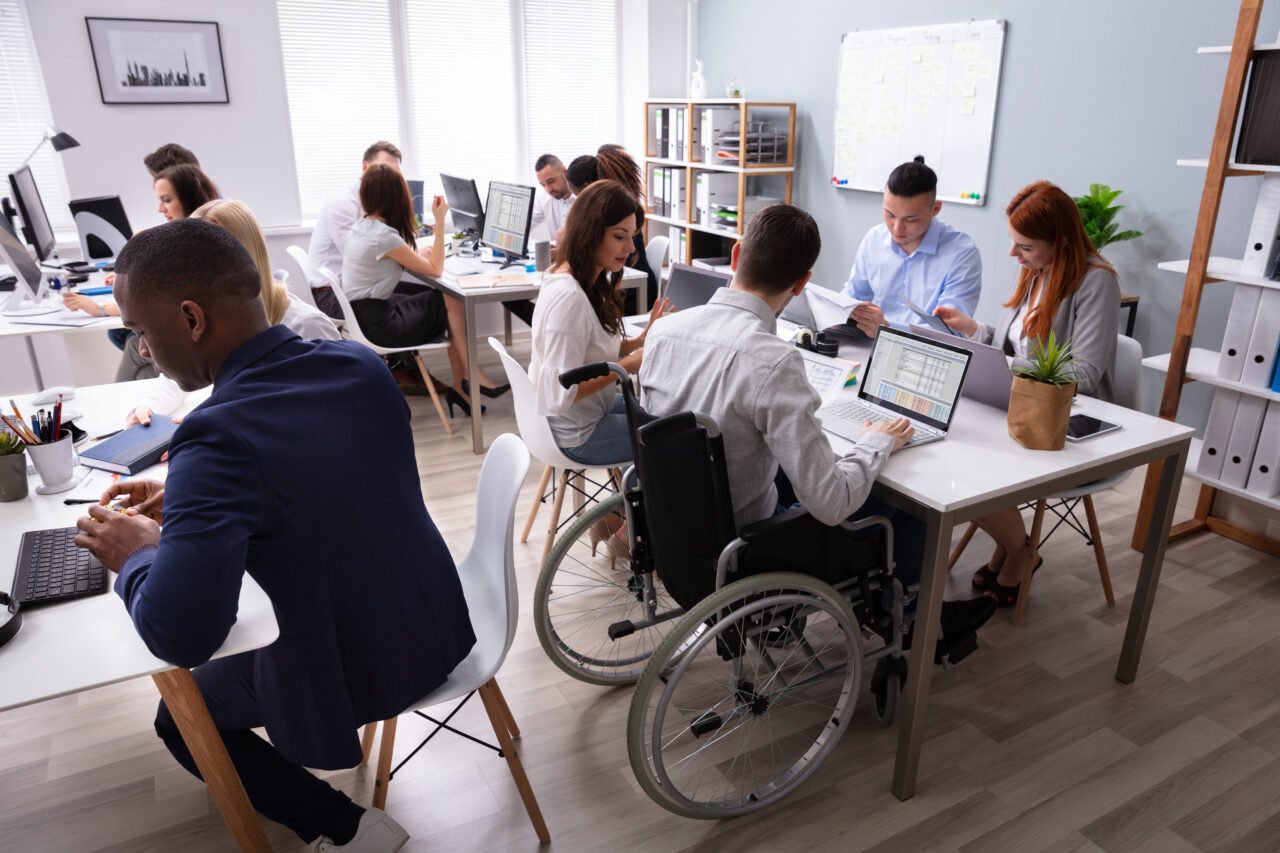 Ludzie pracujący przy biurkach w nowoczesnym biurze, mężczyzna na wózku inwalidzkim przy komputerze.