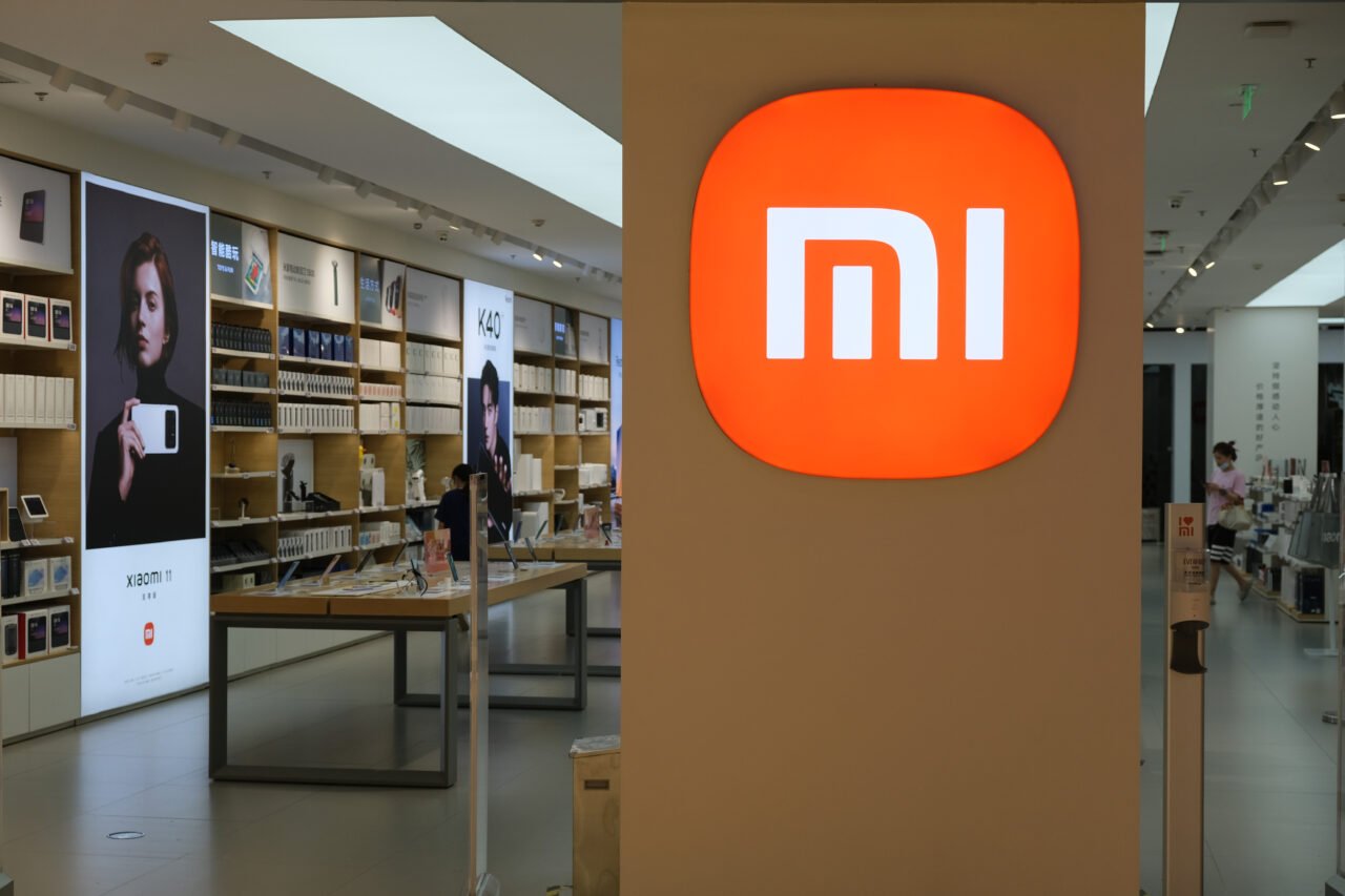 Wnętrze sklepu Xiaomi z dużym, podświetlanym logo Xiaomi na pierwszym planie. W tle półki z produktami i reklamy promujące urządzenia.