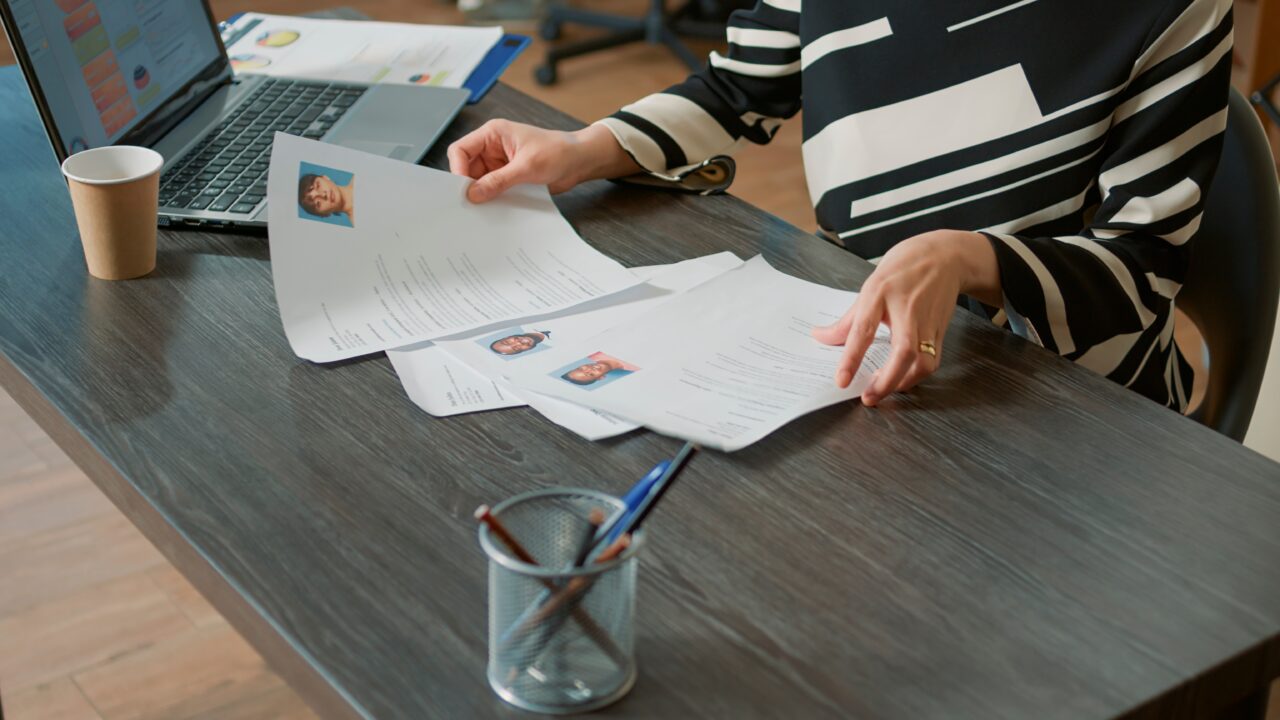 Osoba przeglądająca CV na biurku obok laptopa i kubka.