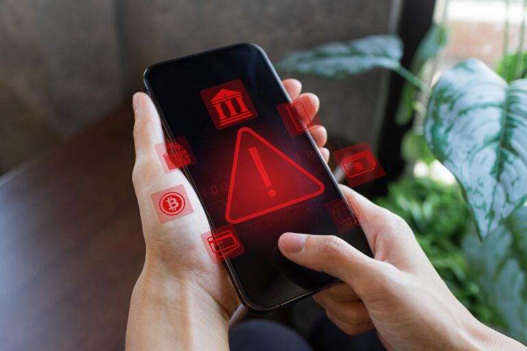 Osoba trzymająca smartfon z wyświetlonym czerwonym symbolem ostrzeżenia oraz ikonami kryptowaluty i bankowości.