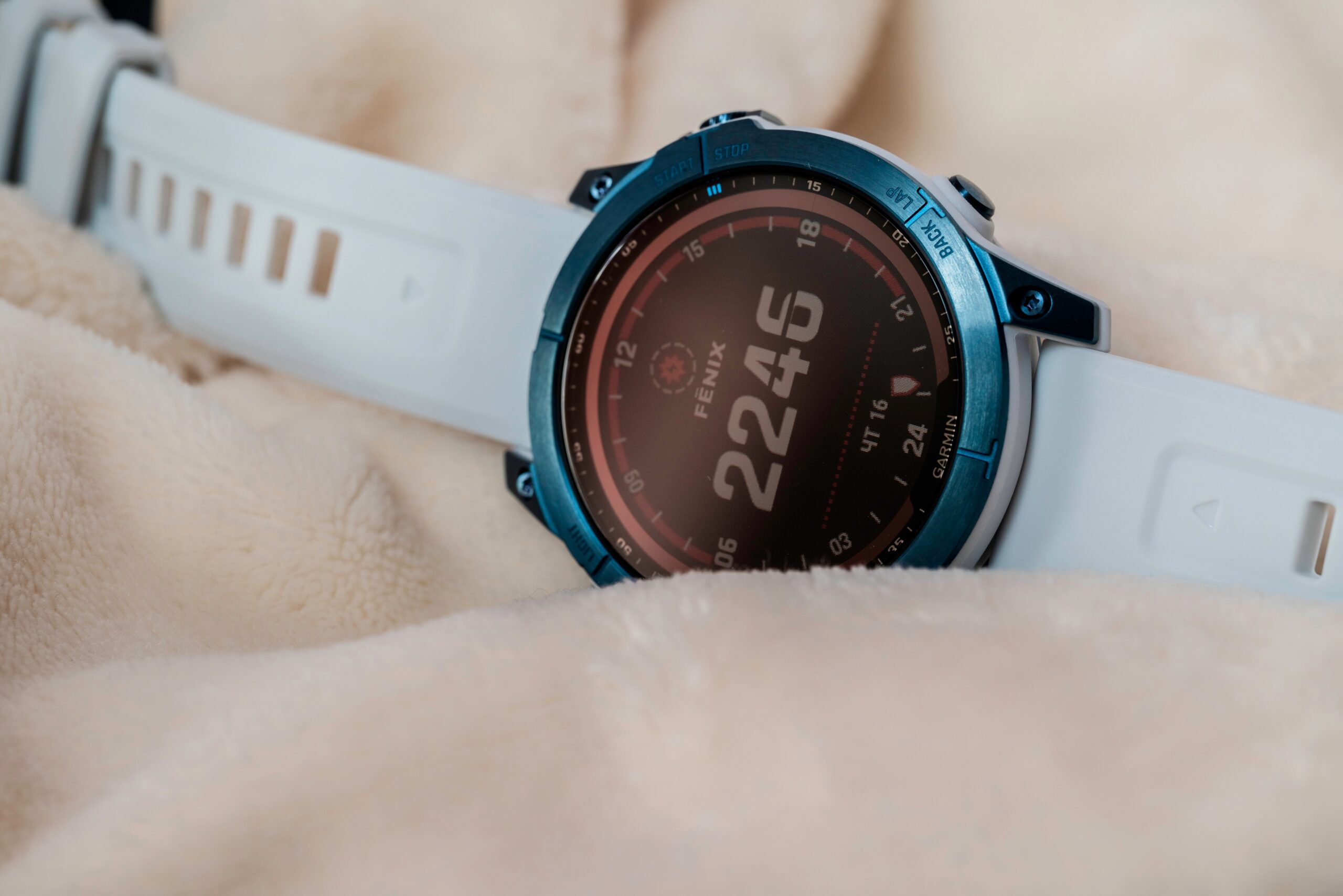 Smartwatch Garmin z serii Fenix z białym paskiem na beżowym kocu.