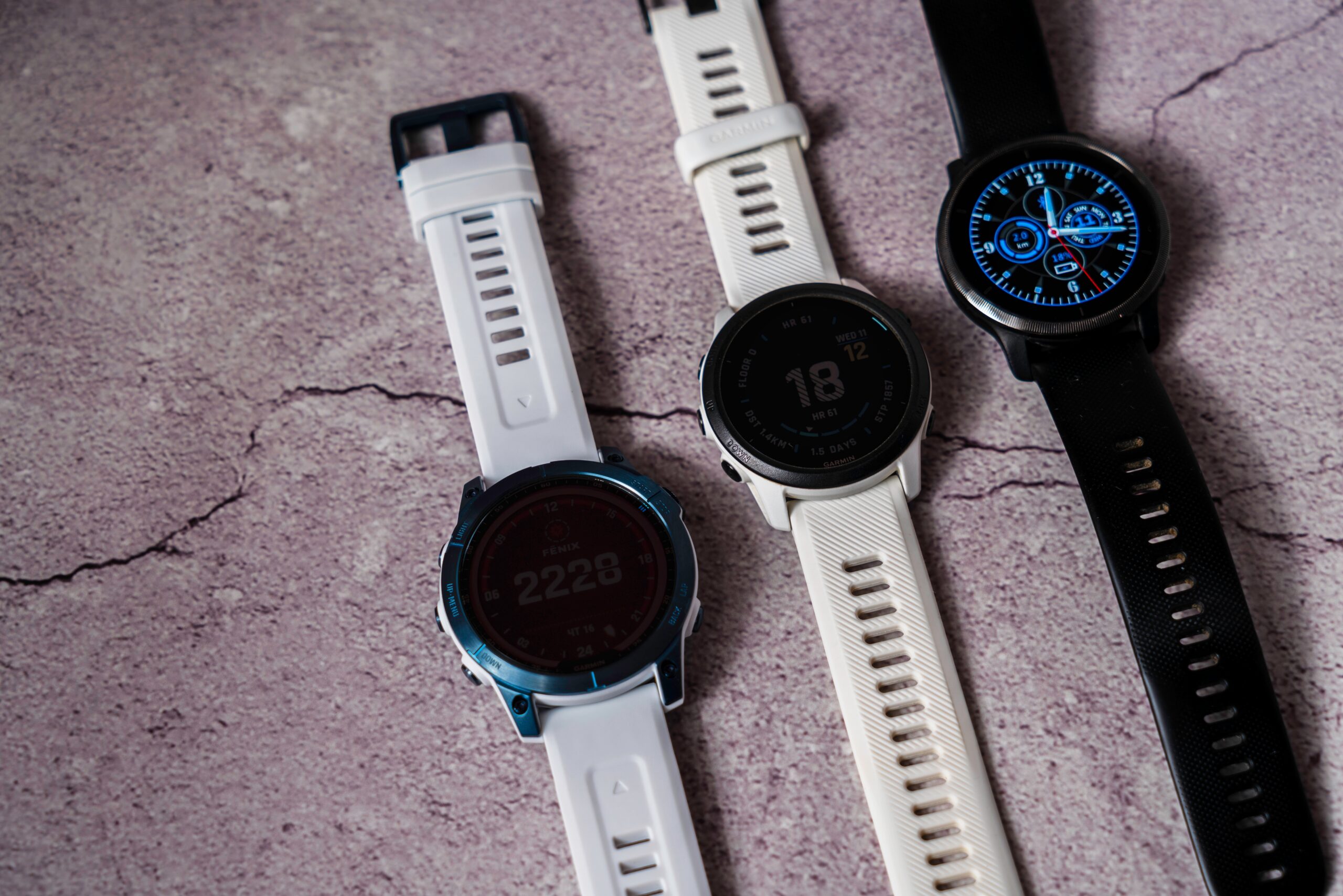 Trzy różne smartwatche na podłożu z betonu, dwa z białymi paskami i jeden z czarnym paskiem.