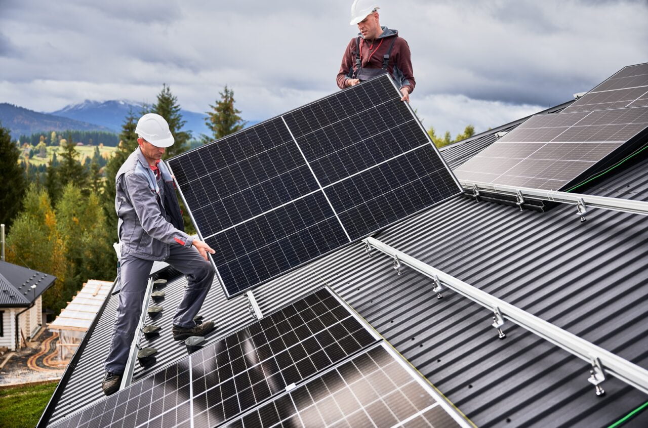 Dwóch pracowników montujących panele słoneczne na dachu budynku w otoczeniu górskiego krajobrazu.