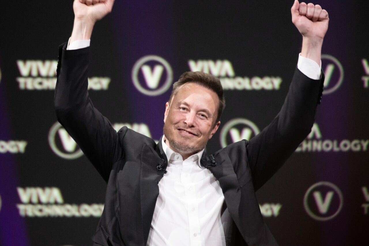 Elon Musk w garniturze unosi ręce w geście triumfu na tle logo Viva Technology.