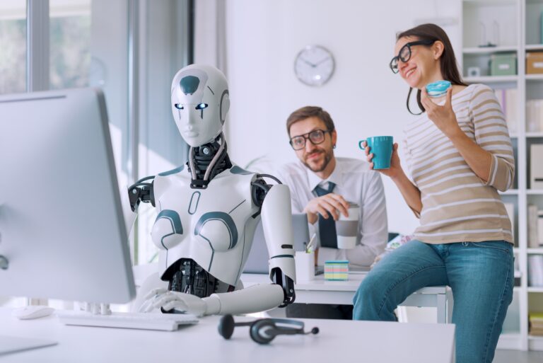 Robot przy komputerze w biurze, przyglądający się i rozmawiający z dwójką ludzi pijących kawę.