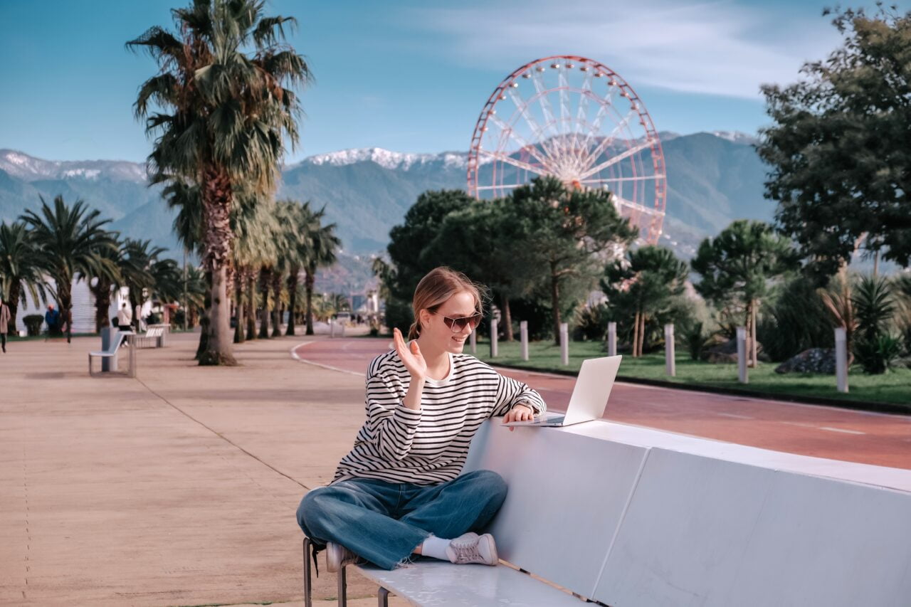 Kobieta w okularach przeciwsłonecznych siedzi na ławce w parku, używając laptopa; w tle widać palmy, alejkę spacerową oraz diabelski młyn i góry.