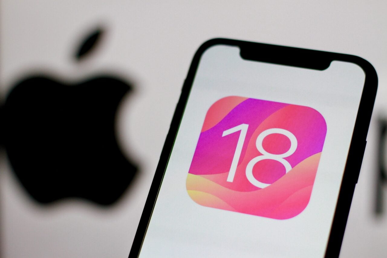 Kolejne nowości w iOS 18 . Telefon iPhone z wyświetlonym logo ios 18 systemu operacyjnego iOS 18, w tle rozmyte logo Apple.