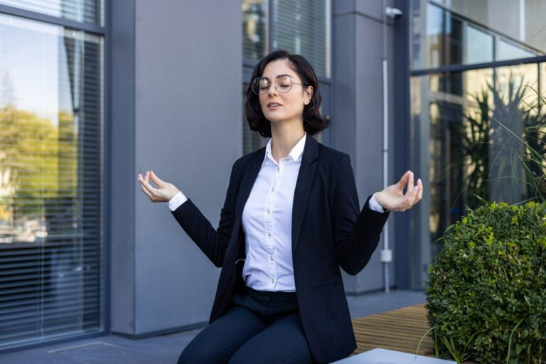 Kobieta w garniturze i okularach medytuje na zewnątrz budynku. 4-dniowy tydzień pracy lub skrócenie czasu przebywania w biurze może pozytywnie wpłynąć na samopoczucie pracowników.