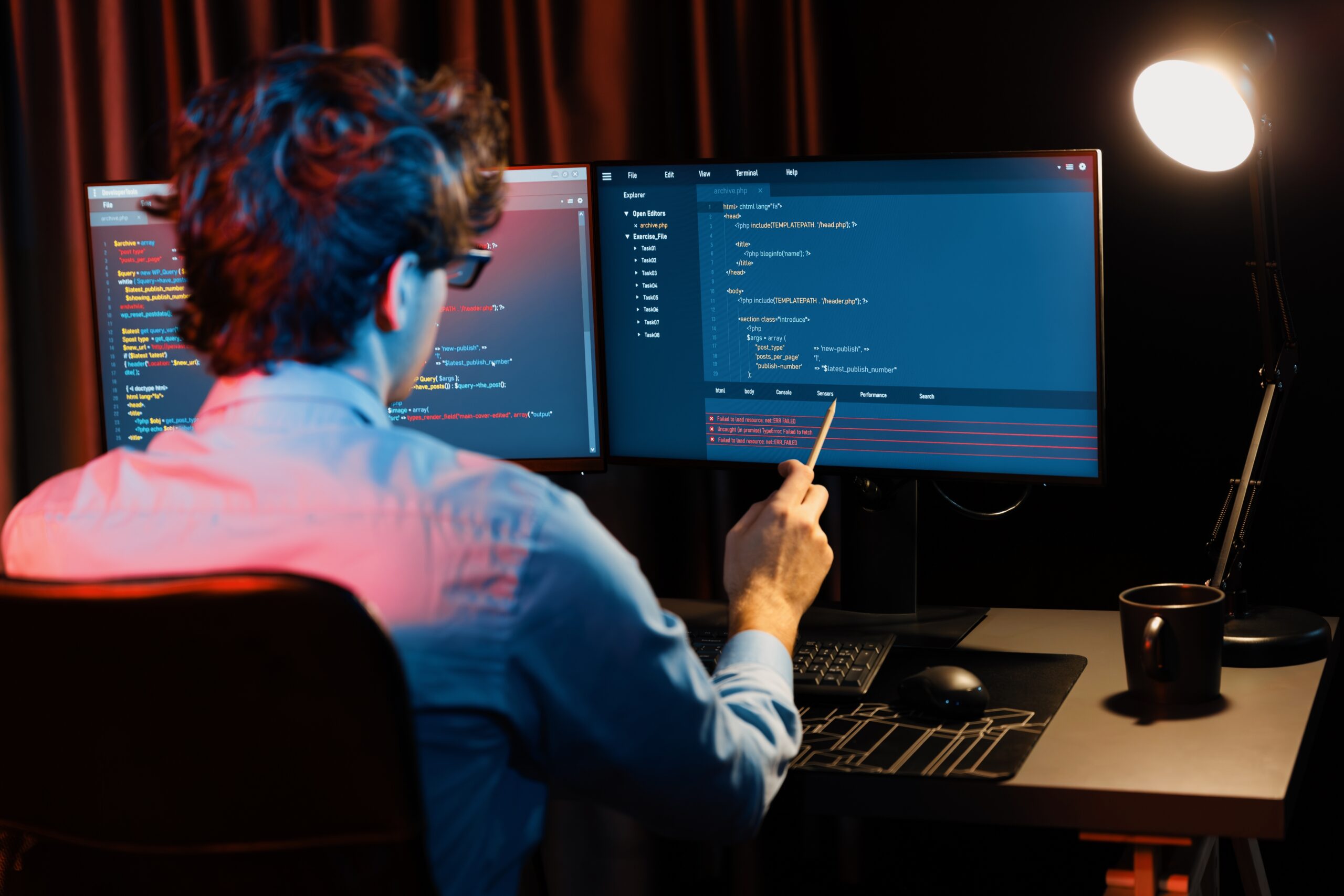 Mężczyzna programista pracujący przy dwóch monitorach komputerowych w zaciemnionym pokoju, wskazuje na kod na ekranie.