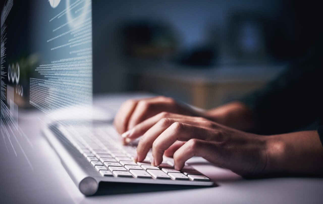 Oszustwo z AI - jak się chronić? Osoba pracująca na laptopie z grafiką kodu komputerowego wyświetlaną na ekranie.