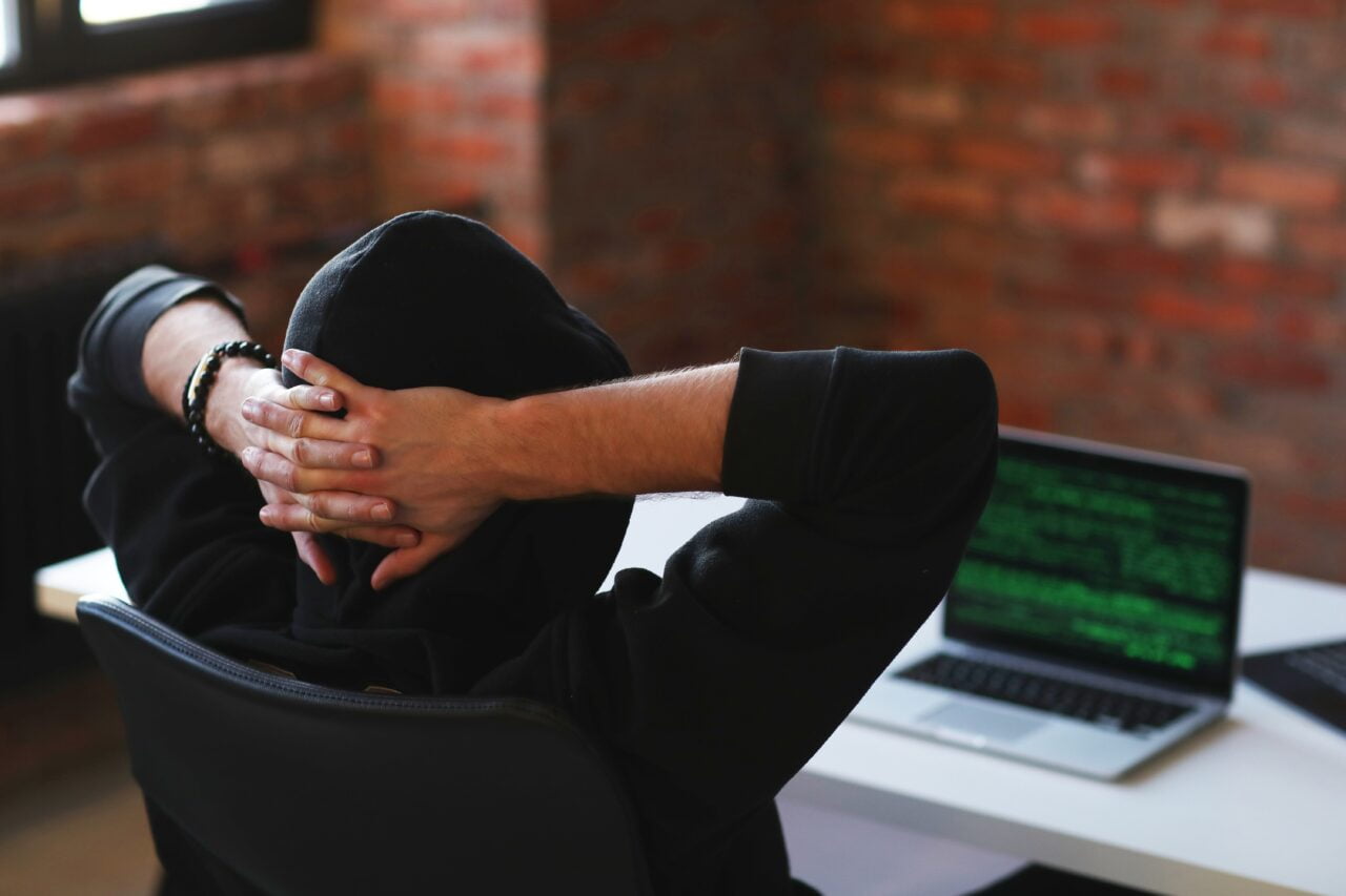 Osoba w czarnej bluzie z kapturem, siedząca tyłem do kamery, opiera głowę na splecionych dłoniach przed laptopem z kodem programistycznym na ekranie, w pomieszczeniu z ceglaną ścianą.
