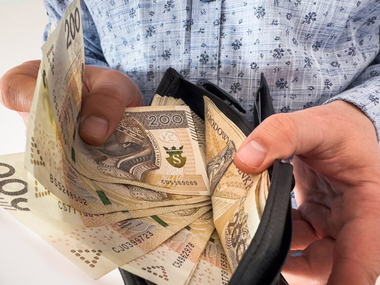 Mężczyzna w jasnoniebieskiej koszuli wyciąga z czarnego portfela polskie banknoty o różnych nominałach.