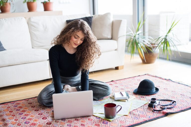 Kobieta z długimi kręconymi włosami siedzi na dywanie, korzystając z laptopa. Obok niej leżą zeszyty, mapa, kapelusz, kubek i aparat fotograficzny. W tle widoczne są sofa i rośliny w doniczkach.