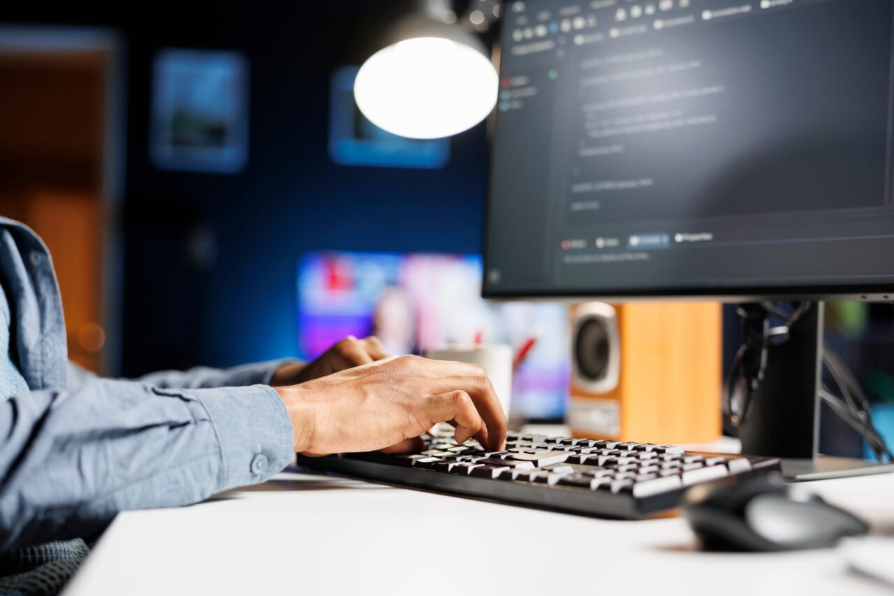 Osoba pracująca przy komputerze, pisząca na klawiaturze w zaciemnionym pokoju.