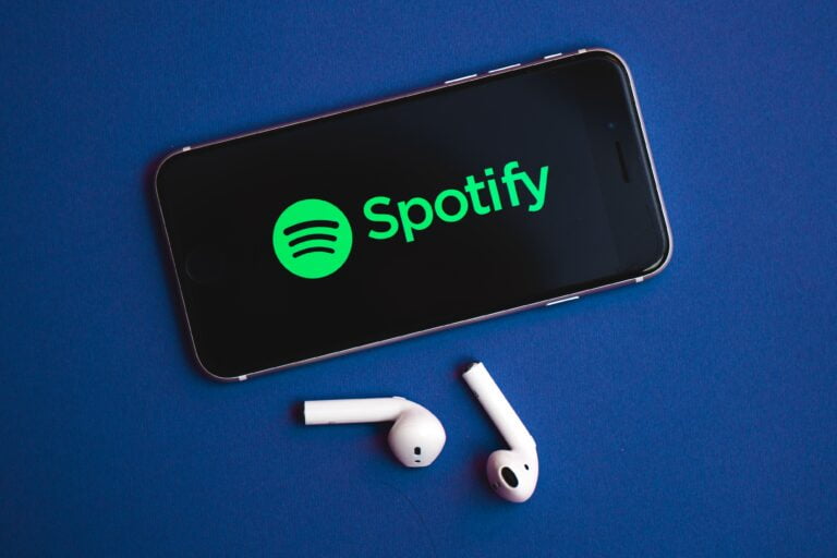 Telefon z otwartą aplikacją Spotify i leżące obok słuchawki bezprzewodowe, na niebieskim tle.